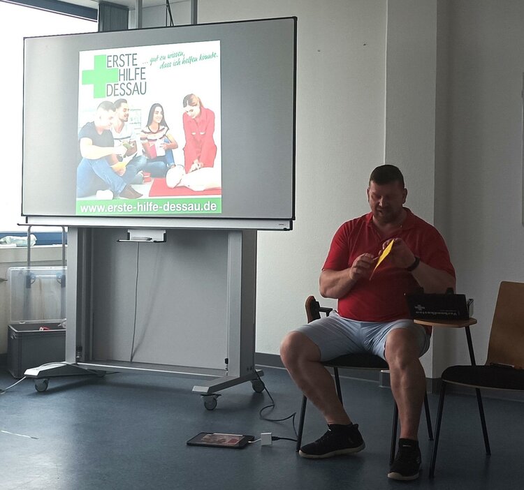 Ein Mann in einem roten Poloshirt und grauen Shorts sitzt auf einem Stuhl und hält ein gelbes Objekt in der Hand. Im Hintergrund ist ein großer Bildschirm mit einer Präsentation zu sehen, auf der mehrere Personen bei einer Erste-Hilfe-Schulung abgebildet sind. Der Bildschirm zeigt auch die Website-Adresse "www.erste-hilfe-dessau.de".