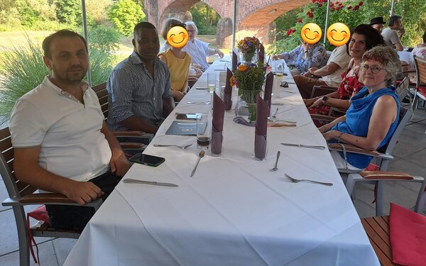 Eine Gruppe von sechs Personen sitzt an einem langen, festlich gedeckten Tisch auf einer Terrasse mit Blick auf einen grünen Garten und historische Backsteinbögen. Auf dem Tisch stehen Blumen, Gläser und Servietten. Einige Personen lächeln, andere haben Emoji-Gesichter, um sie zu anonymisieren.