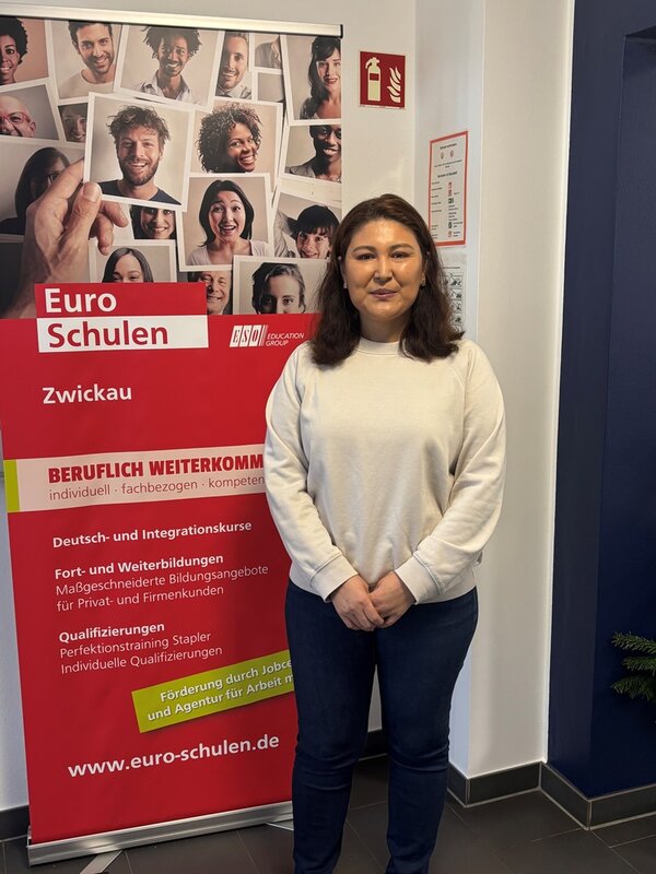 Eine engagierte weibliche Lehrerin steht vor einem roten Werbebanner der Euro-Schulen Zwickau. Sie trägt einen weißen Pullover und eine dunkelblaue Hose. Das Banner hinter ihr zeigt unscharfe Porträtfotos und enthält Informationen zu beruflicher Weiterbildung, Deutsch- und Integrationskursen sowie Qualifizierungen. Die Lehrerin steht in einem hellen Raum mit grauen Fliesenboden und einer blauen Wand rechts. Neben ihr ist ein Feuerlöscher-Symbol an der Wand angebracht.