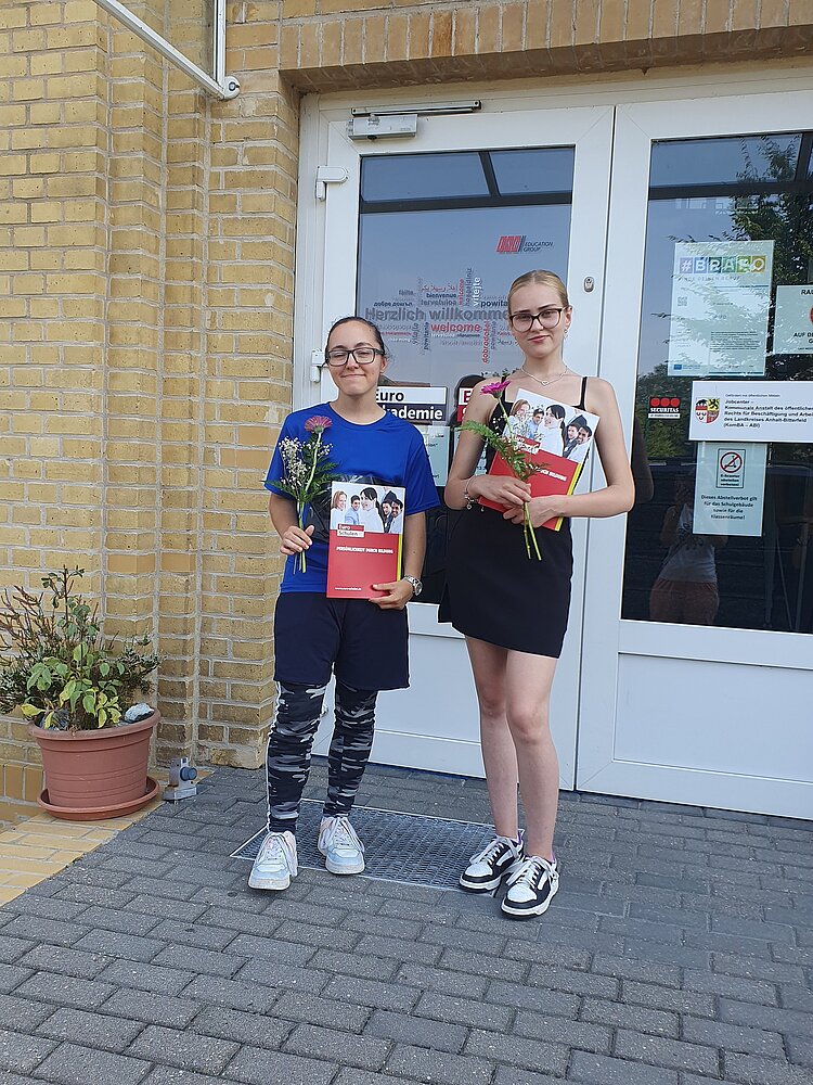 Geschafft! Zwei Absolventinnen stehen vor der Tür zu den Euro-Schulen. Sie halten die Prüfungszeugnisse in einer Mappe der Euro-Schulen sowie kleine Blumensträußchen mit pinken Gerbera in der Hand.