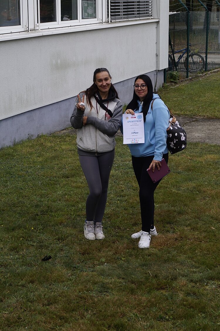 Rückblick Sportfest 2024 Zwei Schülerinnen stehe auf einer Wiese vor dem Gebäude der Euro-Schulen Hoyerswerda und zeigen ihre Urkunden.