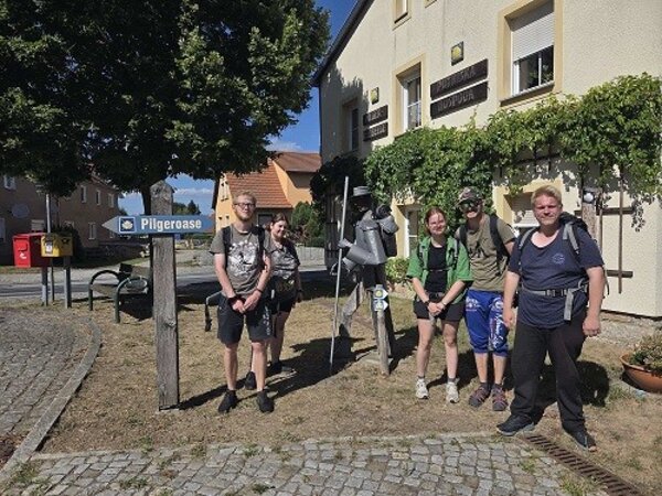 Eine Gruppe von Pilgern steht vor einem Gebäude mit einem Schild, das auf die 'Pilger-Oase' hinweist. Die Personen tragen Rucksäcke und lächeln in die Kamera. Im Hintergrund sind Bäume und ein gepflasterter Weg zu sehen.