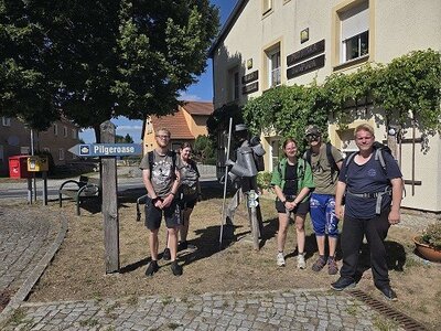 Eine Gruppe von Pilgern steht vor einem Gebäude mit einem Schild, das auf die 'Pilger-Oase' hinweist. Die Personen tragen Rucksäcke und lächeln in die Kamera. Im Hintergrund sind Bäume und ein gepflasterter Weg zu sehen.
