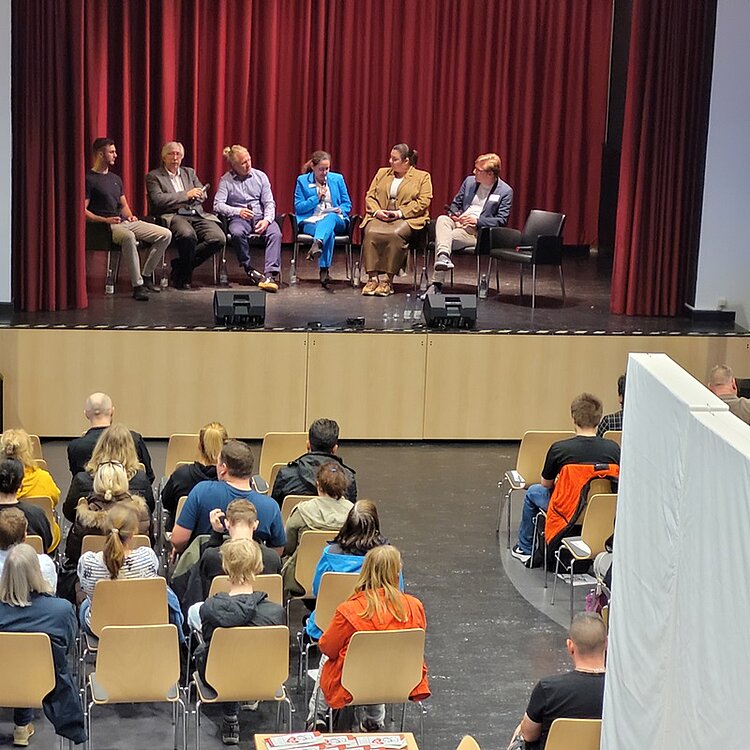 Lebhafte Podiumsdiskussion und Markt der Möglichkeiten Teilnehmende sitzen im Halbkreis auf der Bühne und diskutieren, das Publikum hört zu.
