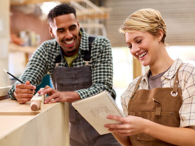Zwei Personen befinden sich in einer Werkstatt. Eine Person hält einen Bleistift und scheint etwas auf einem Stück Holz zu markieren oder zu messen, während die andere Person ein Notizbuch hält und offenbar Notizen macht. Beide Personen tragen Schürzen, was darauf hindeutet, dass sie handwerkliche Arbeiten ausführen. Im Hintergrund sind verschiedene Werkstattelemente wie Werkzeuge und Materialien zu sehen.