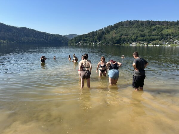 Eine Gruppe junger Menschen steht bis zu den Knien in einem Badesee; im Hintergrund sind bewaldete Hügel zu sehen.