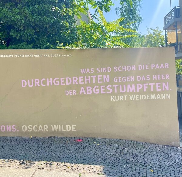 Auf einer hellen Mauer steht ein Zitat in großen, pastellfarbenen Buchstaben: 'WAS SIND SCHON DIE PAAR DURCHGEDREHTEN GEGEN DAS HEER DER ABGESTUMPF­TEN. KURT WEIDEMANN'. Darüber befindet sich in kleiner Schrift ein weiteres Zitat: 'OBSESSIVE PEOPLE MAKE GREAT ART. SUSAN SONTAG'. Im unteren Bereich ist ein weiteres Zitatfragment in Rosa zu erkennen, das mit 'OSCAR WILDE' endet. Im Vordergrund verläuft ein gepflasterter Gehweg, im Hintergrund wachsen Bäume und Sträucher, rechts ist ein Balkon zu sehen.