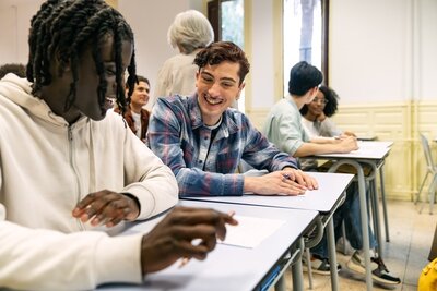 Mehrere junge Menschen sitzen in einem Klassenraum an Schultischen. Im Vordergrund unterhalten sich zwei Personen und lächeln einander an. Sie tragen legere Kleidung und halten Stifte in der Hand. Im Hintergrund sind weitere Personen an Tischen zu sehen, eine ältere Person steht und blickt in den Raum. Die Wände sind hell gestrichen, große Fenster lassen Tageslicht herein.