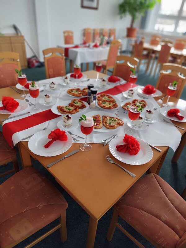 Ein festlich gedeckter Tisch mit Herz-Pizzen, roten Servietten in Blumenform und dekorativen Getränken. Die Farbkombination aus Rot und Weiß verleiht dem Tisch eine elegante und romantische Atmosphäre.