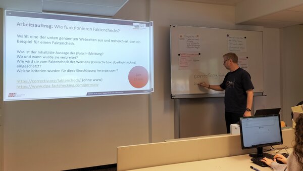 Auf die Wand vorne in einem Klassenzimmer wird ein Bild mit Verhaltenspunkten bei Fake News projiziert.