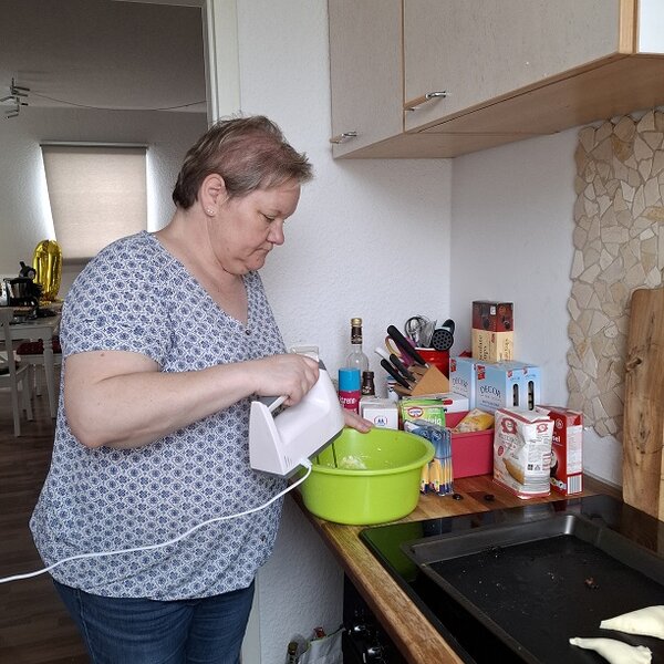 Mitarbeiterin steht in ihrer Küche und bearbeitet den Teig mit dem Handrührgerät in einer hellgrünen Rührschüssel. 