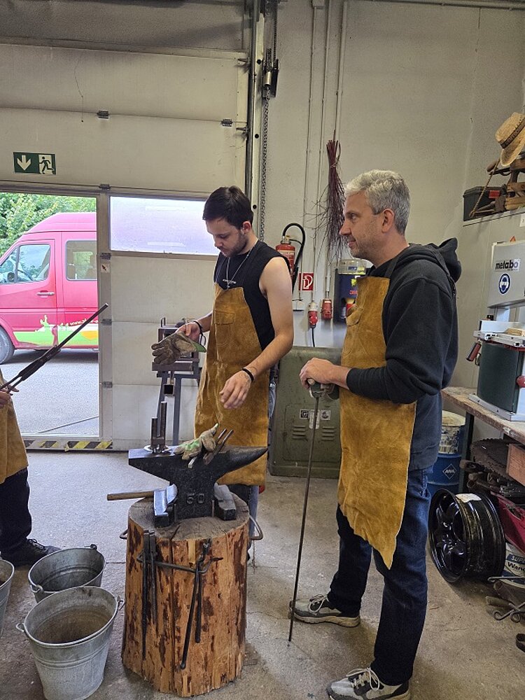 Zwei Personen in Schutzschürzen arbeiten in einer Schmiede, im Vordergrund ein Amboss auf einem Holzblock.