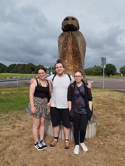 Abschlussfeier der Azubis Die drei Auszubildenden posieren gemeinsam für ein Erinnerungsfoto vor einer hölzernen großen Skulptur, die auf einem Feld an der Ecke einer Kreuzung steht..