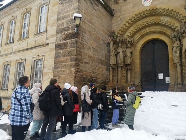 Vor einem historischen Sandsteingebäude mit einem großen, verzierten Rundbogenportal stehen mehrere Personen in Winterkleidung auf verschneitem Boden. Die Fassade zeigt Reliefs und Figuren neben dem Eingang. Die Menschen tragen Mäntel, Mützen und Rucksäcke und blicken in Richtung der Tür. Rechts und links des Portals sind Skulpturen in Nischen zu sehen. Ein Laternenmast ist an der Wand befestigt.