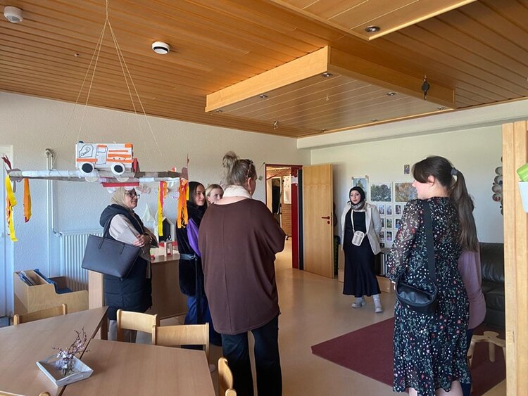 Mehrere Teilnehmende des Berufssprachkurses stehen in einem Raum mit Holzdecke und hellem Mobiliar. An der Decke hängt eine Dekoration in Form eines Fahrzeugs mit bunten Bändern. Im Hintergrund sind eine offene Tür, eine Pinnwand mit Fotos oder Zeichnungen und ein Heizkörper zu sehen. Die Teilnehmenden tragen unterschiedliche Kleidung, darunter ein gemustertes Kleid und eine Jacke mit Kapuze. Die Gesichter sind unkenntlich gemacht.