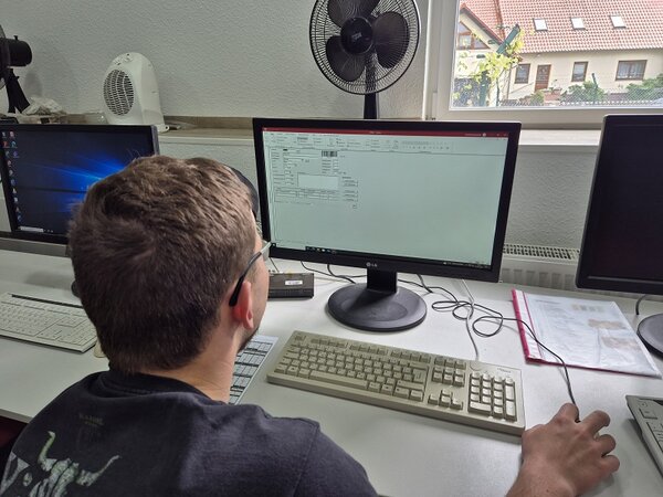 Ein Auszubildender sitzt am Computer und arbeitet mit einer Lagerverwaltungssoftware. Im Hintergrund ist ein Fenster mit Blick nach draußen zu sehen.