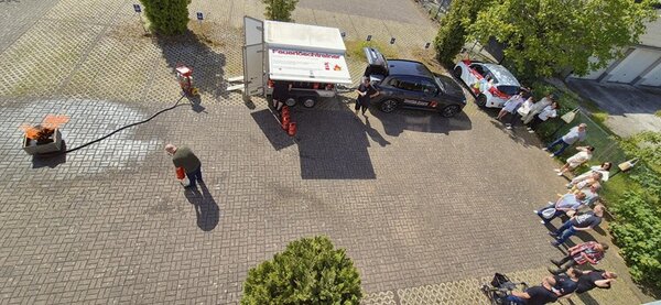 Luftaufnahme eines gepflasterten Parkplatzes mit mehreren geparkten Autos und einer Gruppe von Menschen, die am Rand stehen. In der Mitte befindet sich ein Anhänger mit auffälliger Beschriftung, der an ein dunkles Auto angehängt ist. Ein Mann steht mit einem Feuerlöscher an einem brennenden Objekt, das auf einer kleinen Plattform platziert ist. Die Szene spielt sich bei Tageslicht ab, Bäume spenden teilweise Schatten.