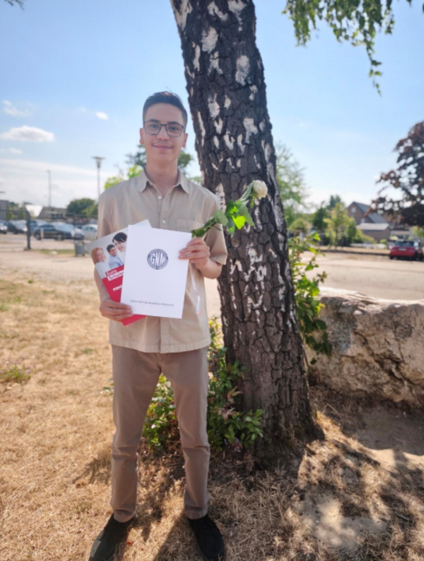 Ein junger Mann mit Brille steht im Freien neben einem Baum und hält eine weiße Rose sowie Unterlagen und eine Broschüre in den Händen. Im Hintergrund sind eine Straße, parkende Autos und Gebäude zu sehen.