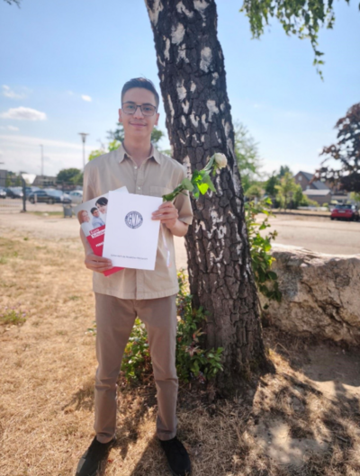 Ein junger Mann mit Brille steht im Freien neben einem Baum und hält eine weiße Rose sowie Unterlagen und eine Broschüre in den Händen. Im Hintergrund sind eine Straße, parkende Autos und Gebäude zu sehen.