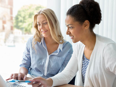 Zwei Frauen sitzen nebeneinander und arbeiten gemeinsam an einem Laptop. Eine Frau trägt ein blaues Hemd und die andere ein weißes Hemd mit einem gestreiften Shirt darunter. Beide konzentrieren sich auf den Bildschirm, während sie etwas auf dem Laptop betrachten oder bearbeiten.