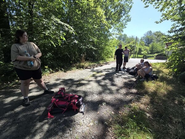 Eine Gruppe von Pilgern macht eine Pause auf einem Waldweg. Einige sitzen auf dem Boden, während andere stehen oder sich unterhalten. Ein roter Rucksack liegt im Vordergrund, und die Szene ist von grünen Bäumen und Sträuchern umgeben.