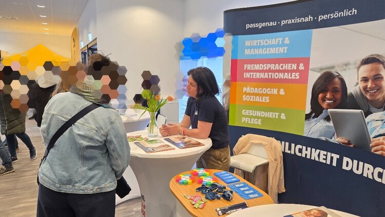 Im Vordergrund steht ein runder Stehtisch mit bunten Werbeartikeln und Flyern. Dahinter unterhält sich eine Mitarbeitende mit zwei Interessentinnen. Eine davon trägt eine helle Jacke. Rechts befindet sich ein Roll-up mit farbigen Textfeldern und einer Abbildung eines Laptops. Im Hintergrund sind weitere Personen und Stände in einem hellen, modernen Raum zu sehen.