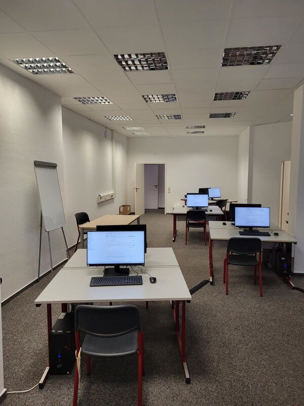 Ein moderner Computerraum mit mehreren Arbeitsplätzen, die mit Monitoren, Tastaturen und Mäusen ausgestattet sind. An der Seite steht ein Whiteboard. Der Raum wirkt ordentlich und funktional.
