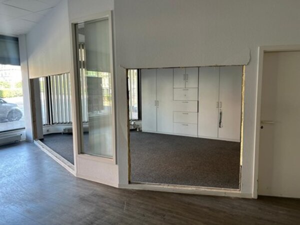 Blick durch eine teilweise entfernte Wand in einen Raum, in dem an der gegenüberliegenden Wand Einbauschränke zu sehen sind. Links gibt es große, bodentiefe Fenster. Draußen ist ein Auto zu sehen.