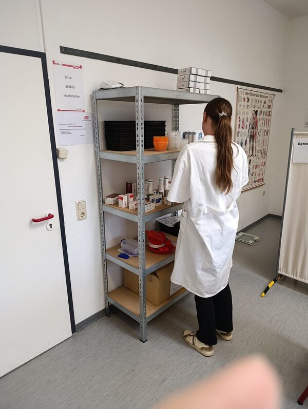 Eine Schülerin in weißem Laborkittel steht vor einem Metallregal mit medizinischen Übungsmaterialien, Medikamentenpackungen und verschiedenen Behältern. An der Wand hängt ein Anatomieposter des menschlichen Körpers.