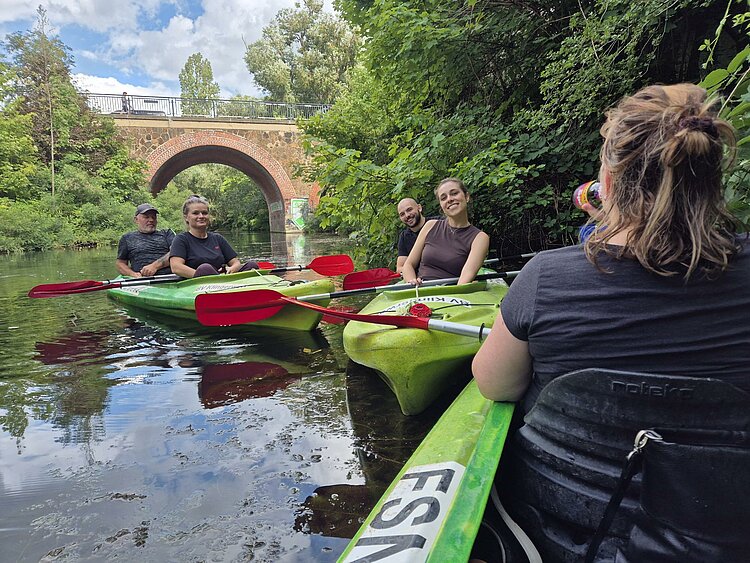 BGM-Sommerferienprogramm an ES und EA Leipzig 2025 Gruppe von Menschen sitzt in grünen Kajaks mit roten Paddeln auf einem Fluss. Im Hintergrund ist eine rote Backsteinbrücke sowie dichtes Grün am Ufer zu sehen.