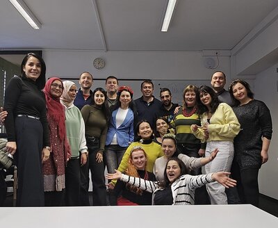 Die Teilnehmenden eines Integrationskurses stehen und sitzen in einem Raum mit weißen Wänden und Decken. Im Hintergrund sind eine Wanduhr, ein Fenster mit Jalousien und eine weiße Tafel zu sehen. Die Personen tragen unterschiedliche, teils farbenfrohe Kleidung, darunter Pullover, Blusen, Jacken und Kopftücher. Einige Personen stehen, andere sitzen oder knien, und vorne streckt ein Kind die Arme weit aus. Die Beleuchtung erfolgt durch lange Deckenleuchten. Alle posieren gut gelaunt für ein Gruppenbild.