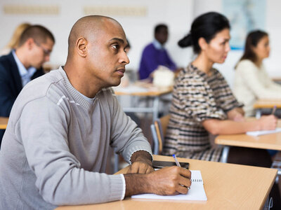 Ein Mann sitzt konzentriert an einem Tisch in einem Klassenzimmer und schreibt in ein Notizbuch. Im Hintergrund sind weitere Personen zu sehen, die ebenfalls an Tischen arbeiten. Die Szene spiegelt eine Lernumgebung wider, die Teil eines Integrationskurses für Menschen mit geringen Lese- und Schreibkenntnissen in lateinischen Schriftzeichen ist.
