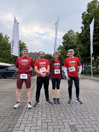 Vier Personen in roten Trikots der Euro Akademie und Sportschuhen stehen auf einem Parkplatz und lächeln in die Kamera.