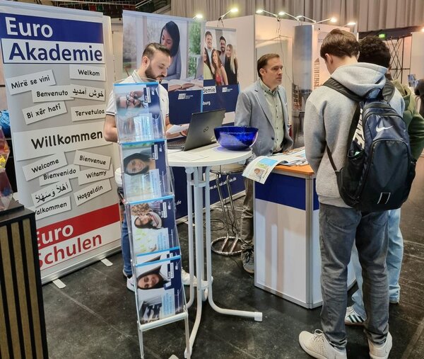 Zwei junge Männer mit Rucksäcken stehen vor dem Stand der Euro Akademie auf dem Karrieretag Oldenburg 2025. Ein Mitarbeiter in einem hellblauen Sakko erklärt ihnen etwas und zeigt Broschüren. Im Vordergrund ist ein Prospektständer mit Infomaterialien zu sehen. Ein weiteres Teammitglied arbeitet an einem Laptop. Der Stand ist mit Bannern gestaltet, auf denen „Euro Akademie“, "Euro-Schulen" und mehrsprachige Begrüßungen stehen.
