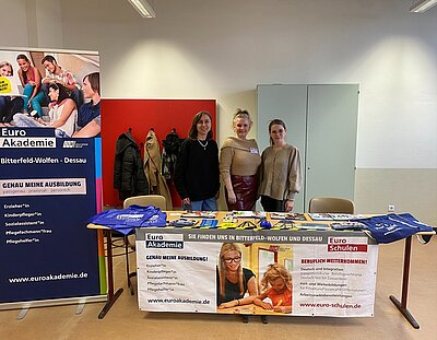 Start unserer Messesaison Drei gute gelaunte Mitarbeiterinnen stehen am Euro Akademie-Messestand, mit großem Roll-Up, Infobroschüren, Jutebeuteln und Stiften auf dem Tisch.