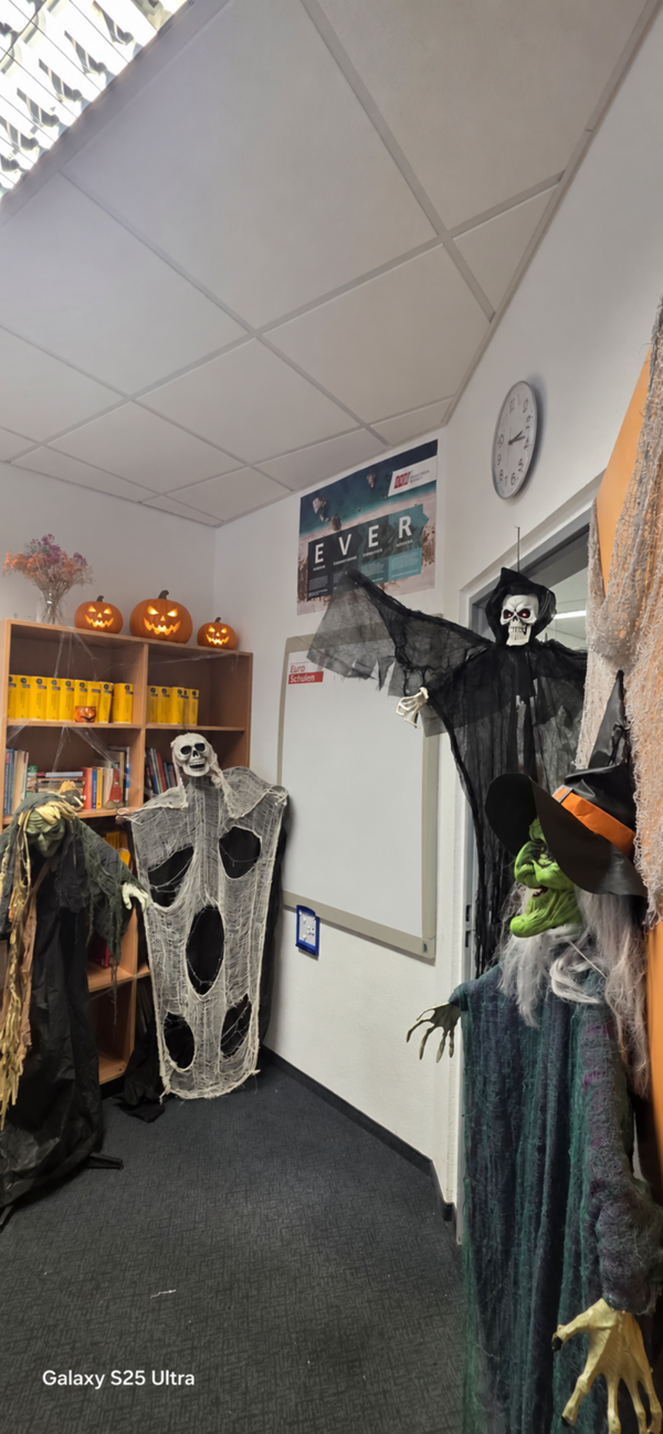 Herbstliche Halloween-Dekoration in einem Innenraum: Mehrere gruselige Figuren wie Skelette, Hexen und ein schwebend wirkender Sensenmann schmücken den Raum. Auf einem Regal stehen leuchtend orangefarbene Kürbisse mit geschnitzten Gesichtern. Die Dekoration verbreitet eine schaurige Halloween-Stimmung.