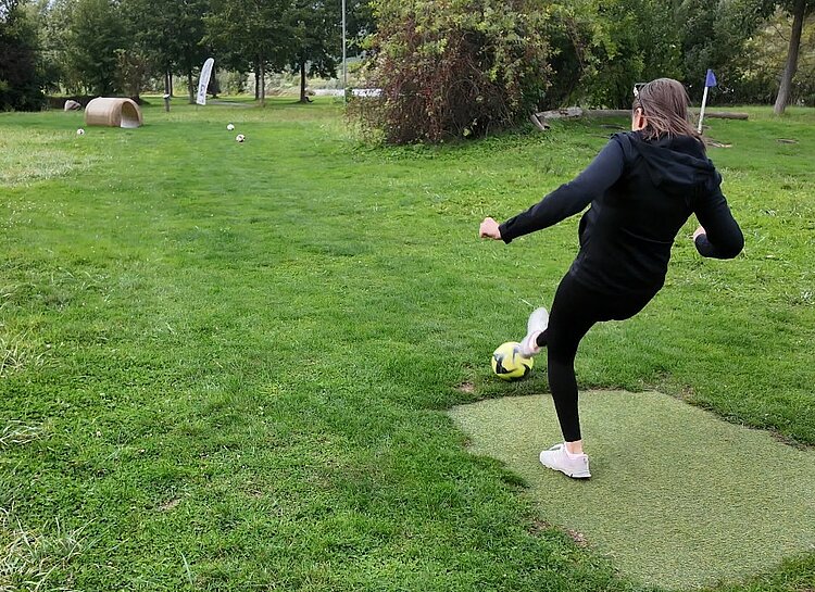 Team-Building beim Fußballgolf Eine Mitarbeitende in schwarzer Kleidung tritt mit ihren weißen Turnschuhen gegen ihren Fußball. Dieser soll wohl durch das ein paar Meter entferne Rohr rollen.