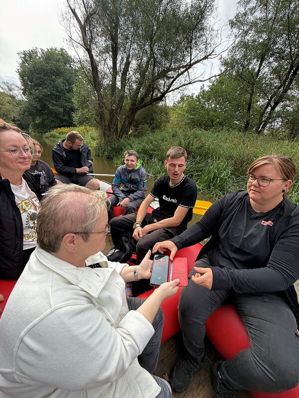 Eine Gruppe von Menschen sitzt in einem roten Schlauchboot auf einem Fluss. Eine Person zeigt einer anderen etwas auf ihrem Smartphone. Die Umgebung ist grün und naturnah.