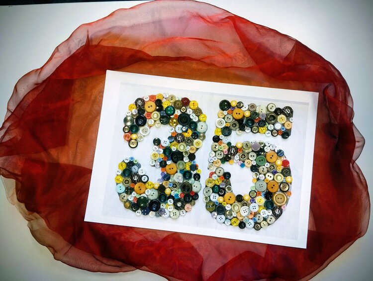 Ein aus bunten Knöpfen gefertigtes Bild der Zahl 35 auf einem weißen Papier liegt auf einem roten Tuch, das wiederum auf einer weißen Fläche liegt.