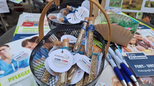 Ein Korb mit kleinen Glasflaschen, die Samen enthalten, steht auf einem Tisch neben Broschüren und Stiften. Die Flaschen tragen Etiketten mit der Aufschrift 'Klimaschule'.