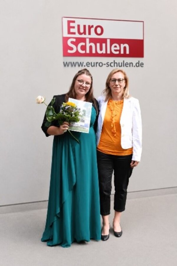 Zwei Frauen stehen lächelnd nebeneinander vor einem Hintergrund mit dem Logo von 'Euro-Schulen' und der Website www.euro-schulen.de. Die Frau links hat ein grünes langes Kleid an und hält einen Blumenstrauß sowie ein Zertifikat in den Händen, während die Frau rechts in einem orangefarbenen Oberteil und einer weißen Jacke gekleidet ist.