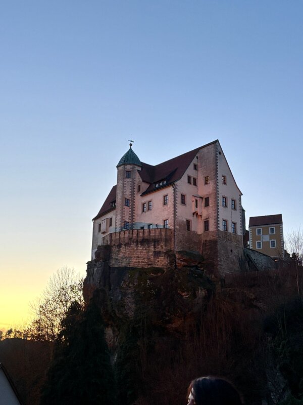Historisches Schloss auf einem Felsen bei Sonnenuntergang, mit beleuchteten Fenstern und klarem Himmel in der Dämmerung.