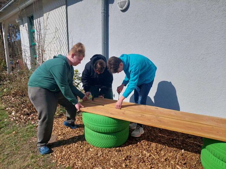 Drei Jugendliche montieren im Außenbereich eine selbst gebaute Sitzbank aus Holzbrettern und grün gestrichenen Reifen.