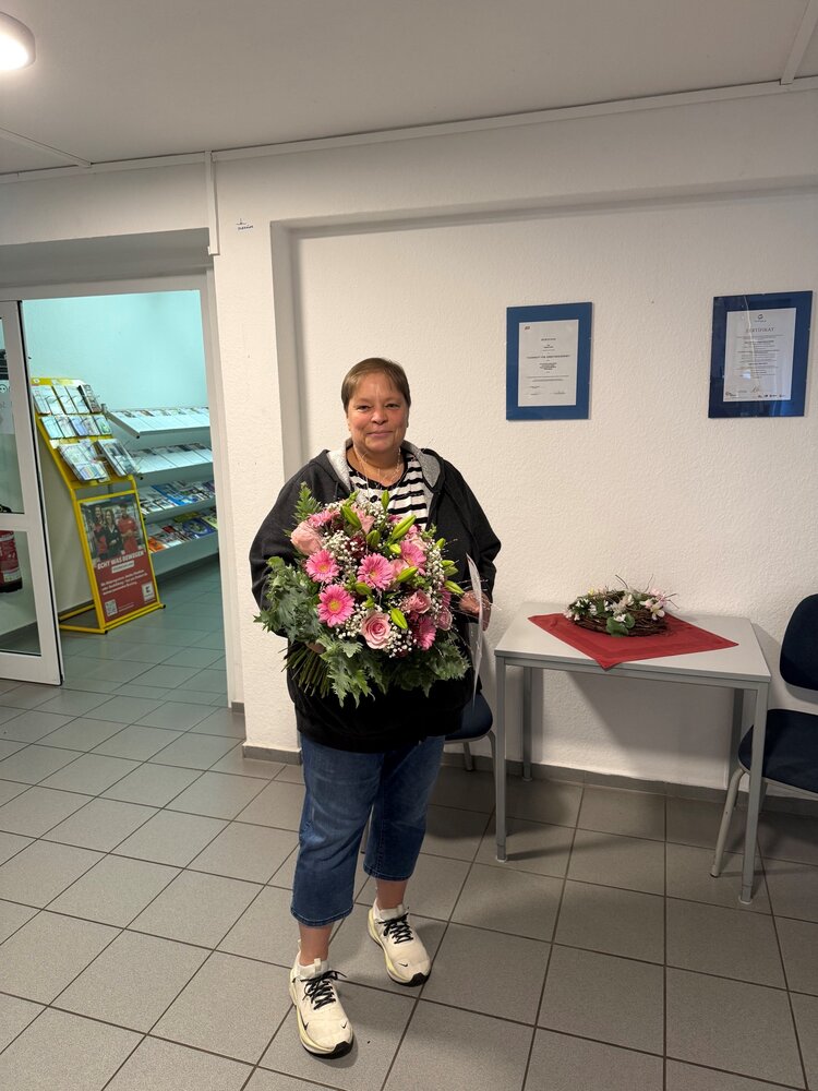 Eine Frau steht mit einem großen Blumenstrauß aus rosa Rosen, Gerbera und weiteren Blumen in den Räumen der Euro-Schulen und blickt lächelnd in die Kamera.