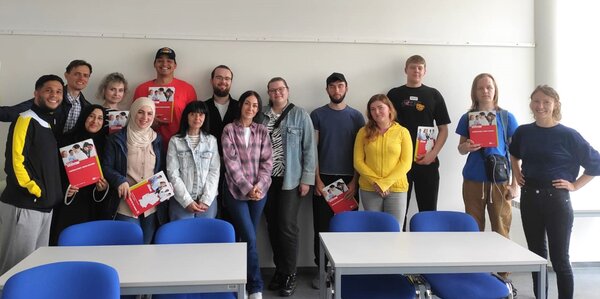 Eine Gruppe von fünfzehn jungen Erwachsenen steht lächelnd in einem Klassenraum vor einer weißen Wand. Einige halten rote Informationsmappen mit dem Titel „Fachwirt/in im Gesundheits- und Sozialwesen“. Vor der Gruppe stehen leere Tische mit blauen Stühlen.
