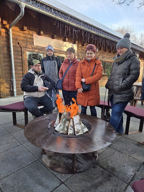 Eine Gruppe von Menschen steht im Freien um eine runde Feuerstelle und wärmt sich an einem offenen Holzfeuer vor einem Holzgebäude