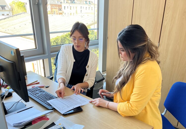 Zwei Frauen sitzen an einem Schreibtisch in einem Büro und besprechen gemeinsam ein Dokument; eine der Frauen zeigt mit dem Finger auf eine Textstelle, während die andere aufmerksam zuhört und einen Stift in der Hand hält.