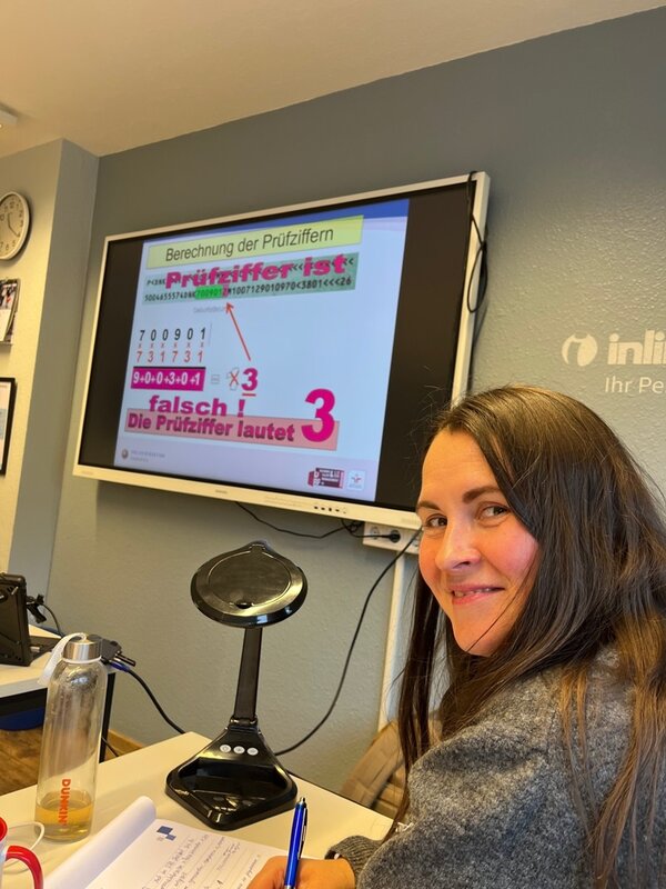 Eine Mitarbeiterin der Euro-Schulen Hannover sitzt am Tisch, macht sich Notizen und lächelt in Richtung Kamera. Hinter ihr läuft auf einem Monitor eine Präsentation zur Ermittlung der Prüfziffer in Ausweisdokumenten, Teil der polizeilichen Schulung zum Thema Dokumentenfälschung. Auf dem Tisch stehen eine Dokumentenkamera, eine Wasserflasche und Schulungsunterlagen.