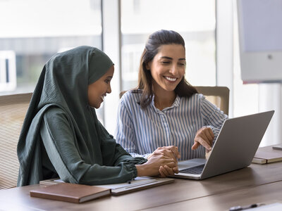 Zwei lächelnde Frauen sitzen gemeinsam an einem Tisch in einem hellen Büro oder Besprechungsraum. Die Frau links trägt ein grünes Oberteil und ein grünes Kopftuch. Die Frau rechts trägt ein gestreiftes Hemd und zeigt auf den Bildschirm eines offenen Laptops, der vor ihnen steht. Beide wirken freundlich und engagiert, während sie sich über den Inhalt des Laptops austauschen. Der Raum ist modern und gut beleuchtet, mit großen Fenstern im Hintergrund.