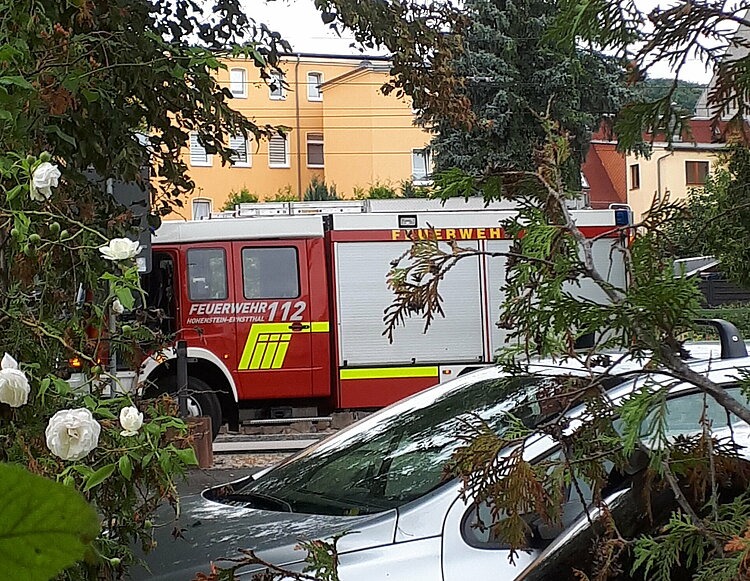 Ein rotes Löschfahrzeug der Feuerwehr mit der Notrufnummer 112 ist teilweise verdeckt hinter Bäumen und parkenden Autos zu sehen.